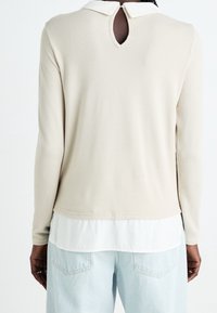 Beige langemouwen top met een witte kraag, met een sleutelgat uitsparing aan de achterkant en gelaagd over een wit shirt, gecombineerd met lichtblauwe jeans.