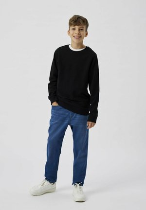 Maglia nera a maniche lunghe con scollo tondo bianco, abbinata a jeans blu e sneakers bianche, in piedi su uno sfondo neutro.