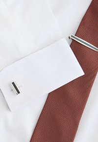 Biała mankiet koszuli z srebrnym spinkiem, w zestawieniu z teksturowanym krawatem w czerwono-brązowym odcieniu, z delikatnym wzorem. Prosty i elegancki design.
