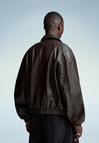 Brown leather bomber jacket with a textured finish, elastic waistband, and cuffed sleeves. Features a classic collar and relaxed fit design.