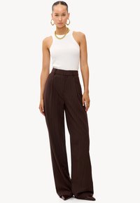 Camiseta de tirantes blanca con ribetes, combinada con pantalones de talle alto marrón con rayas finas. Los pantalones tienen un ajuste holgado y pliegues delanteros sutiles. Accesorios dorados.