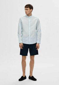 Camicia button-up azzurro chiaro con colletto a banda, abbinata a pantaloni navy e scarpe nere slip-on; tessuto liscio e design informale.