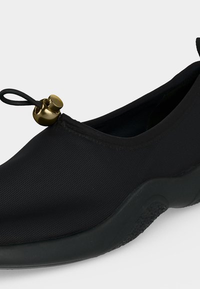 Μαύρο παπούτσι τύπου slip-on με υφασμένη λαστιχένια σόλα και χρυσό κουμπί σε μαύρο ελαστικό κορδόνι στην μπροστινή άνοιγμα για ρύθμιση.