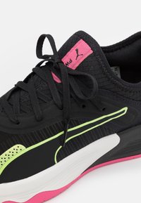 Svart atletisk sneakers med neongröna accenter, vit mellansula, rosa yttersula, svarta snören, och en rosa etikett med en Puma-logotyp på tungan.