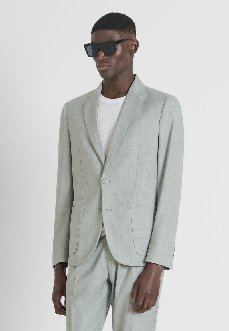 Blazer grigio chiaro e pantaloni abbinati; tessuto testurizzato con un motivo sottile; chiusura a un bottone; abbinato a una maglietta bianca a coste e occhiali da sole neri.
