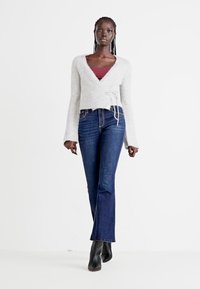 Grau, Wickel-Style Pullover mit langen Ärmeln und Bindedetail, kombiniert mit dunkelblauen Schlagjeans und schwarzen Stiefeletten.
