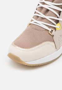 Beige sportovní boty s síťovinovým svrškem, zlatými akcenty a bílými tkaničkami. Hladká gumová podrážka s texturovaným vzorem pro lepší přilnavost.