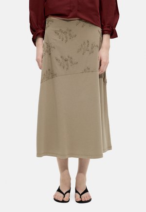 Beżowa spódnica midi z kwiatowym haftem na górnej części, noszona z bordową bluzką i czarnymi sandałami na odkryte palce na jednolitym tle.