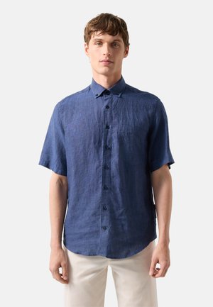 Mann trägt ein blaues kurzärmeliges Hemd mit Kragen und beige Hose vor einem einfarbigen hellen Hintergrund.