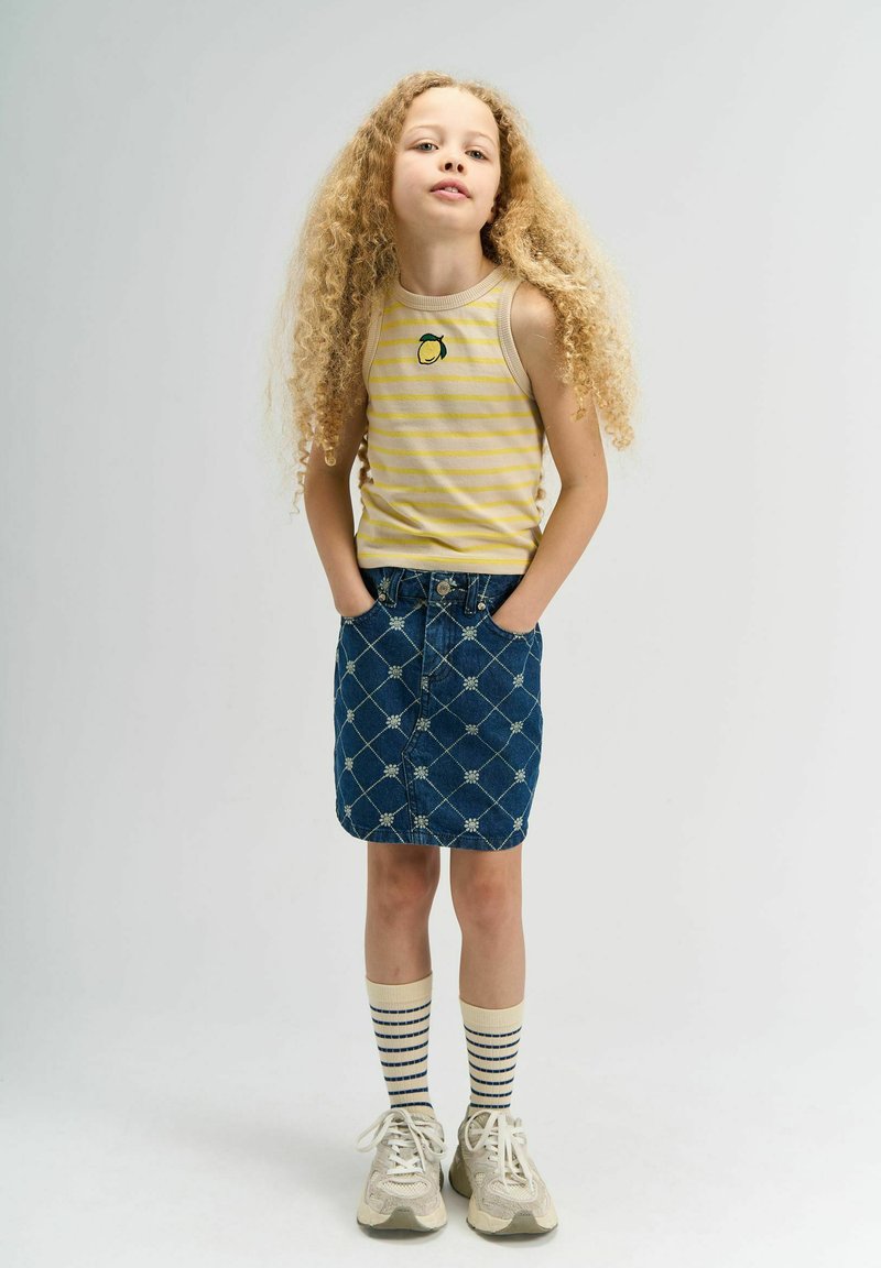 Débardeur jaune rayé avec broderie de citron, jupe en denim bleu avec motif en losange et fleurs blanches, chaussettes crème avec rayures bleu marine, baskets beiges.