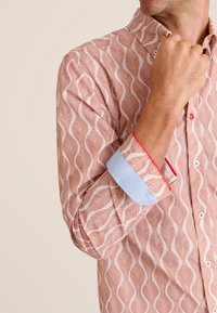 Lange mouw shirt in perzik met wit bladerenpatroon, contrasterende lichtblauwe manchetten en rode afwerking. Katoenen stof met een kraag met knopen.