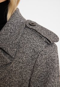 Grijze en zwarte houndstooth-gepatroneerde jas met een gestructureerde kraag, voorzien van een zwarte knop op de schouder en een gestructureerde stof.