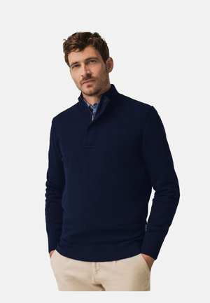 Marineblauwe pullover met een kraag met een kwartrits, geribbelde manchetten en zoom. Zachte, gebreide textuur met een gelaagde uitstraling over een overhemd met knopen.