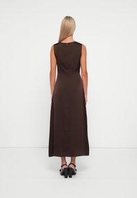 Robe maxi sans manches marron avec un corsage ajusté et une jupe fluide. Tissu lisse, éclat subtil et fermeture éclair au dos. Sandales à talons épais.