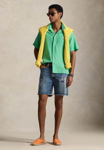 Moški, oblečen v zeleno srajco s kratkimi rokavi, rumen pulover, prevrnjen čez ramena, kratke hlače iz popravljenega jeansa, oranžne natikače in sončna očala.