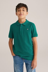 Zöld poloing, texturált anyagból készült, gallérral, három gombbal és egy kis hímzett logóval a mellen. Farmernadrággal viselve.