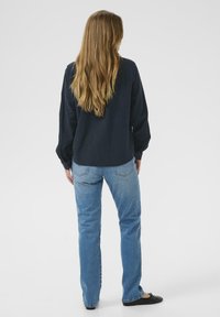 Marineblauw lange mouwen shirt met gathered cuffs, gecombineerd met lichtblauwe straight-leg jeans en zwarte platte schoenen. Het haar is lang en golvend.