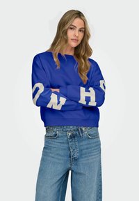 Sweat-shirt bleu marine avec lettrage crème sur les manches, encolure ronde et poignets côtelés. Associé à un jean taille haute bleu clair.