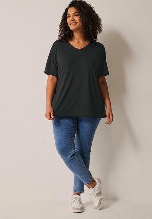 Femme aux cheveux bouclés portant un haut noir ample col en V, un jean bleu et des baskets blanches, debout contre un mur clair uni.