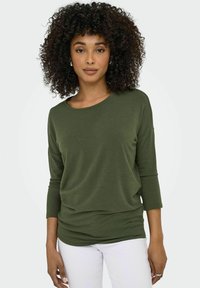 Maglietta a maniche lunghe verde oliva in un morbido tessuto testurizzato, con scollatura rotonda e design drappeggiato, abbinata a pantaloni bianchi.