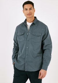 Temno zeleni srajčni gumbasti shirt, izdelan iz teksturirane tkanine. Ima dva žepa na prsih, koničast ovratnik in dolge rokave.