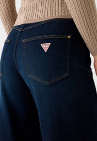 Jeans en denim bleu foncé à coupe large, avec un logo Guess triangulaire rouge et blanc sur la poche arrière et des détails de coutures visibles.
