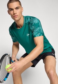 Türkisfarbene Sportshirt mit strukturierten, gemusterten Ärmeln, kombiniert mit schwarzen Shorts. Das Model hält einen Tennisschläger und einen Ball.