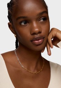 Złoto-srebrny łańcuszek z wydłużonymi owalnymi ogniwami. Modelka nosi kremowy top w prążki i pozują z palcami opartymi na brodzie.