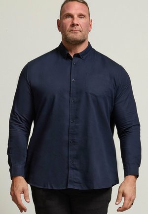 Man standing facing forward, wearing a dark blue long-sleeve button-up shirt with a chest pocket and black pants against a plain background.