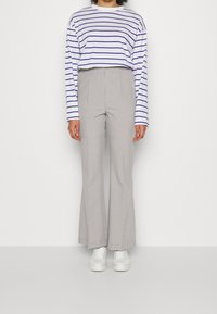 Persona vestita con una camicia bianca a maniche lunghe con righe orizzontali blu, pantaloni larghi grigio chiaro e sneakers bianche, in piedi contro un muro bianco.