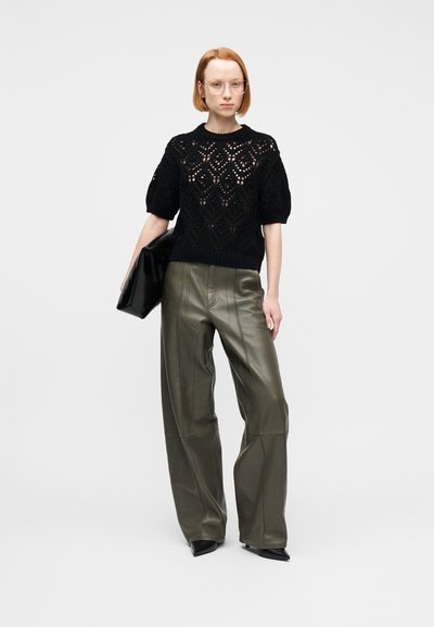 Femeie cu păr roșcat, scurt, purtând ochelari transparenti, pulover negru tricotat cu mâneci scurte, pantaloni largi de piele olive, pantofi negri cu vârf ascuțit, ținând o plic negru.