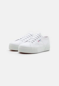 Hvite kanvas sneakers med rund tå, gummisåle og hevet teksturert grep. Funksjoner inkluderer metalløyler og subtil logo detaljer.