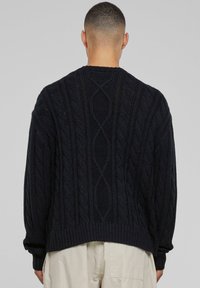 Pull en tricot noir à mailles torsadées avec une coupe décontractée, présentant des poignets et un ourlet côtelés. Motif texturé distinctif dans le dos.