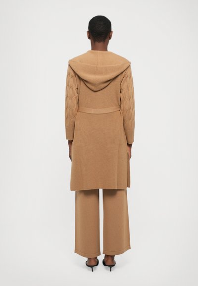 Μακρύ μπεζ καρφίτσα με κουκούλα, μανίκια με καλωδίωση, ζώνη στη μέση και πλευρικές σχισμές, συνδυασμένο με μπεζ παντελόνι με φαρδιά πόδια και μαύρες γόβες.