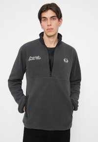 Temno siva flis pulover s polovičnim zadrgo na ovratniku, vezenim besedilom "Originals Sports Club" in logotipom na prsih. Ima stranske žepe.