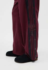 Pantalon de survêtement Adidas bordeaux avec bandes latérales noires, boutons-pression et écusson logo, porté avec des chaussures noires sans lacets.