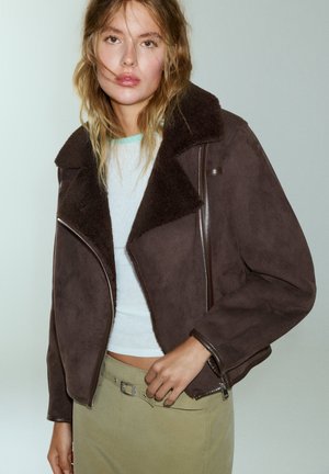 Le modèle porte une veste marron foncé à col en peau lainée sur un haut blanc et une jupe kaki, et pose devant un fond clair.