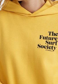 Żółta bluza z kapturem z miękkiego materiału z czarnym nadrukowanym tekstem, który brzmi "Future Surf Society" oraz logo marki.