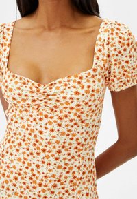 Robe à motif floral crème avec des fleurs orange, décolleté carré, manches courtes bouffantes et tissu texturé. Silhouette ajustée.