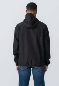 Sort letvægtsjakke med hætte, elastiske manchetter og løstsiddende pasform, vist fra bageste vinkel. Matchet med blå denimjeans.
