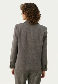 Femme aux cheveux bouclés portant un costume texturé gris, vue de dos sur un fond clair uni.