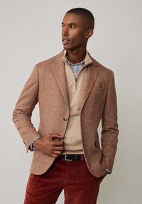 Brauner Hahnentritt-Blazer mit Revers, getragen über einem beigen Pullover mit Reißverschluss und einem blauen Streifenhemd, kombiniert mit roten Cordhosen.
