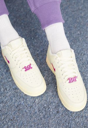 Voeten met crèmekleurige Nike-sneakers met roze swooshes en bijpassende veterlabels, witte sokken en paarse broek op een blauw tapijt.