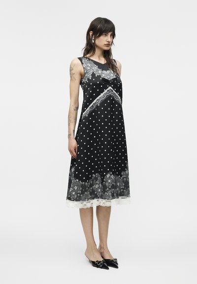 Γυναίκα που φοράει μαύρο μίντι φόρεμα χωρίς μανίκια με λευκές πουά, λεπτομέρειες από δαντέλα και μαύρες μυτερές γόβες, στέκεται μπροστά σε μονόχρωμο φόντο.
