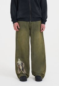 Olivové široké džíny s jemným grafickým motivem křídel a textovým detailem; mají vybledlou texturu a dvě zadní kapsy.