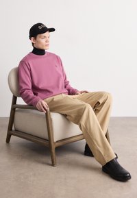 Felpa rosa, dolcevita nero, pantaloni beige e scarpe nere; seduto su una sedia imbottita chiara con struttura in legno.