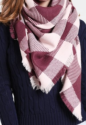 Femme portant un pull en tricot torsadé bleu marine et une grande écharpe à carreaux rose et bordeaux avec des franges, enroulée autour de son cou.