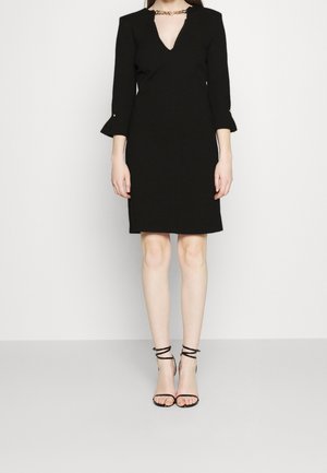 Femme portant une robe noire longueur genou avec des manches trois-quarts et un décolleté en V profond, associée à des talons noirs à bride cheville.