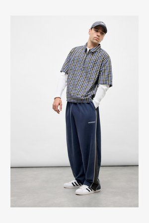 Short-sleeve plaid shirt in blue and yellow, layered over a white long-sleeve T-shirt. Loose navy sweatpants with white side stripes, white sneakers.