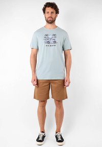 Camisa de algodón de color azul claro con una impresión gráfica de un vehículo y palmeras. Pantalones cortos marrones, de tejido texturizado, y zapatillas negras con suelas blancas.